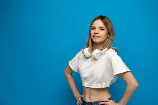 Portrait of beautiful blonde young woman with a white headphones on a shoulder on blue background