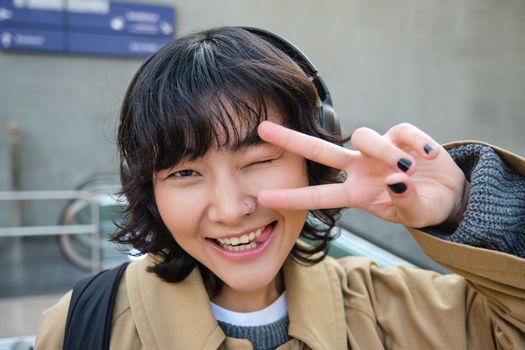 Head portrait of korean girl standing on street, showing peace sign, wearing headphones, listening music and smiling.