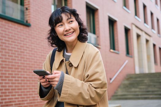 Happy korean girl walks on street, listens music in wireless earphones and holds smartphone, picks song in playlist while standing outdoor near building, reading message.