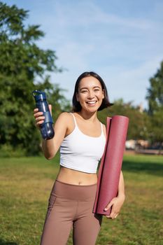 Portrait of happy asian girl, fitness woman giving you water bottle to drink after workout, standing with rubber mat for exercises in park.