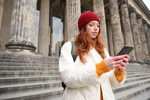 Portrait of young urban girl with backpack, using smartphone, walking around city, looking at map on mobile app, checking direction.