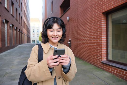 Portrait of young korean woman walking around city with backpack and headphones, listens music, looks at smartphone, uses phone application on streets.