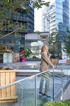 Vertical shot of corporate woman in beige suit standing outside on street, drinks morning coffee takeaway before work.