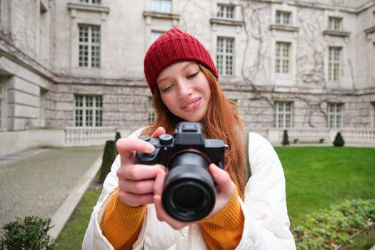 Happy redhead girl tourist, takes photos, photographer with professional camera walks around city and captures beautiful pictures.