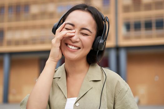 Portrait of beautiful asian woman in headphones, listening music on street of city centre, smiling happily.