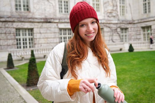 Portrait of happy girl traveler, drinking hot tea from thermos while walking and exploring city. Tourist with flask smiling at camera.