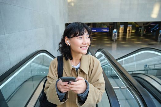 Portrait of cute smiling korean girl, goes up escalator, listens music in wireless earphones and uses mobile phone, holds smartphone.