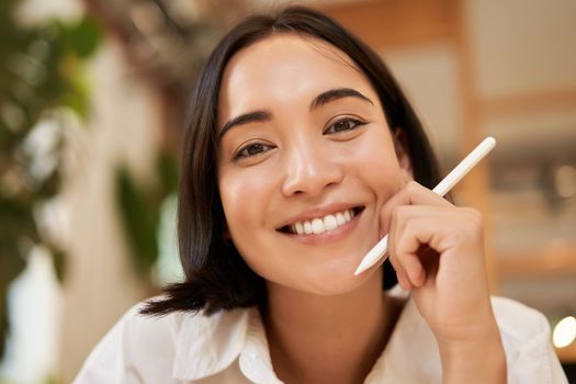 Close up portrait of smiling brunette asian woman, sitting with graphic pen and looking happy, drawing on digital tablet.