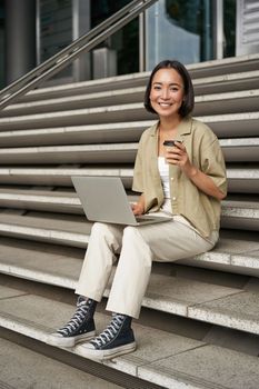 Vertical shot of smiling girl student, asian woman sits on stairs of university campus and drinks coffee, does her homework on laptop.