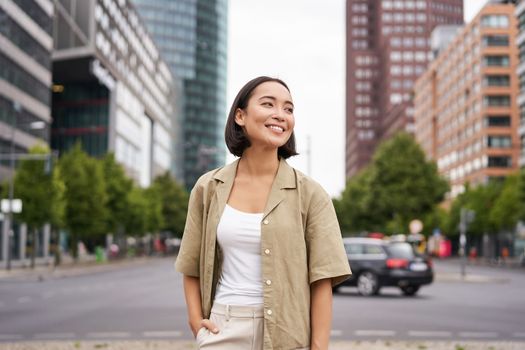 Outdoor shot of young smiling asian woman, standing on street in daytime, looking around, posing happy.