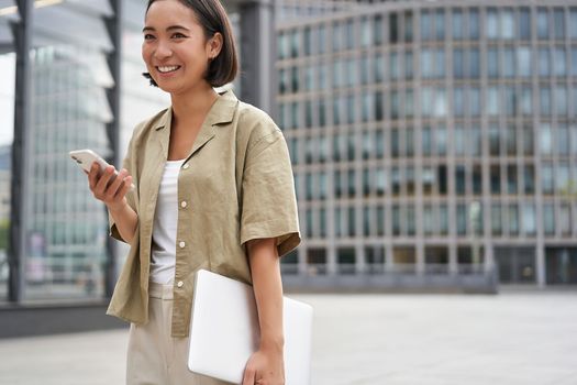 Portrait of asian girl with laptop, going to work or uni, holding smartphone and smiling, walking on street.