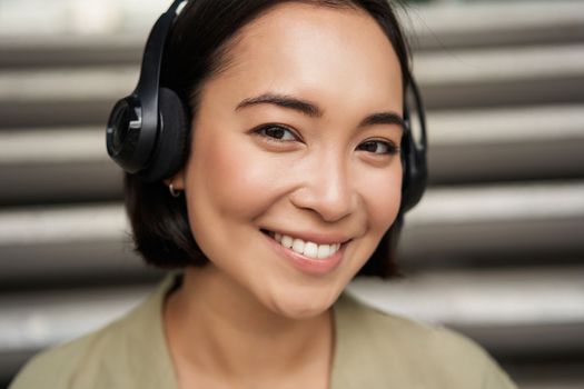 Close up portrait of smiling asian girl in headphones, listens to music outdoors, looking happy. People concept