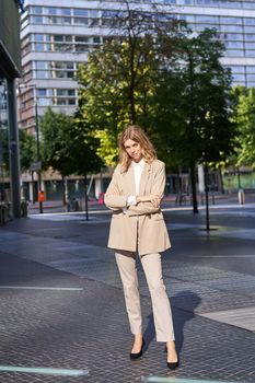 Beautiful businesswoman in beige suit, standing near office buildings on street, posing with confident, relaxed face expression.