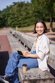 Portrait of young artist, asian girl drawing in digital tablet with graphic pen, sitting outdoors in park, getting inspiration.