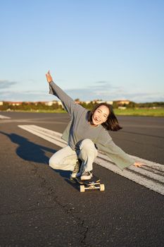 Carefree skater girl on her skateboard, riding longboard on an empty road, holding hands sideways and laughing.