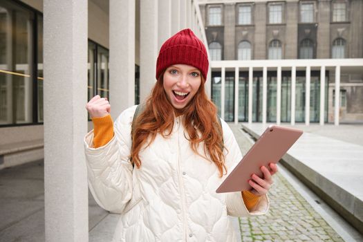 Happy redhead girl in red hat, walks around city with digital tablet, connects to public internet wifi and looks for route, looks at map on her gadget.
