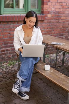 Vertical shot of asian woman working in outdoor cafe, sitting with laptop on bench and drinking coffee. Copy space