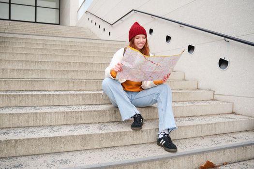 Young smiling redhead girl, tourist sits on stairs outdoors with city paper map, looking for direction, traveller backpacker explores city and looks for sightseeing.