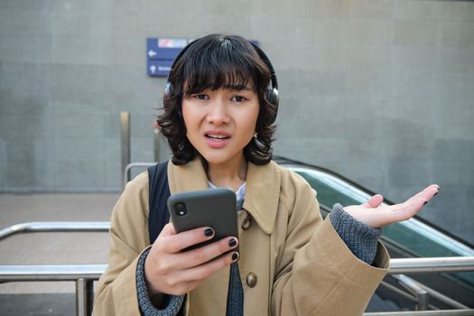 Close up portrait of puzzled, complicated korean girl in headphones, frowns and looks unsure, stands with smartphone and looks at camera while shrugs on street.