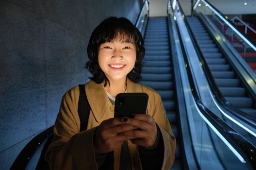 Happy smiling young woman, standing on escalator, going down, holding smartphone in both hands, chatting on mobile phone app.