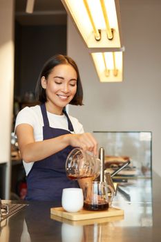 Vertical shot of smiling girl working as barista, prepare pour over, making filter coffee batch brew, standing at counter in cafe in apron.