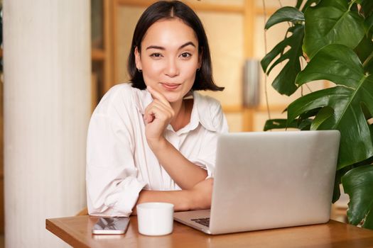 Confident young stylish woman with laptop, sitting in cafe and working, freelancer in co-working space with cup of coffee.