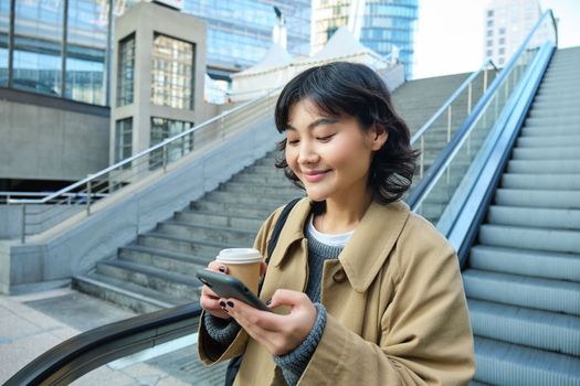 Young stylish girl with cup of coffee, drinks cappuccino to go, goes down escalator and looks at mobile phone, chats, reads message on smartphone.