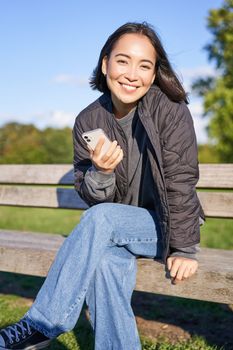 Cute young woman with smartphone in hands, sitting on bench and smiling, using mobile phone, waiting for someone in park.