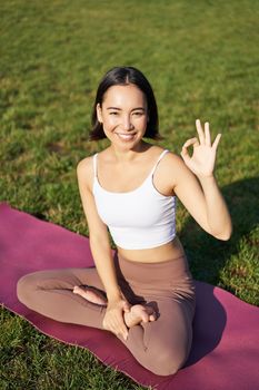 Portrait of asian woman sitting on rubber mat, meditating, showing okay sign and looking at camera, doing yoga.