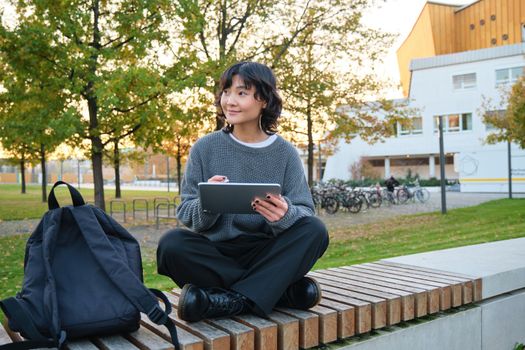 Young girl, korean artist or art student sits in park with digital tablet, draws with graphic pen, scatches a design or project, looks around.