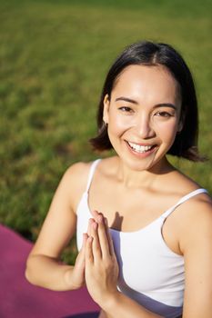 Vertical shot of young asian woman doing yoga, practice mindfulness, smiling and looking relaxed, sitting on rubber mat in park.