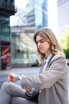 Portrait of office worker in suit, woman looking at watch with disappointed face, reading upsetting message, sitting outdoors.