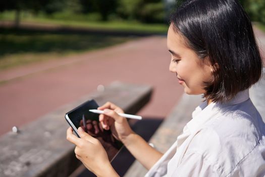 Portrait of young asian woman drawing on fresh air in park, sitting with graphic tablet and digital pen, smiling happy.