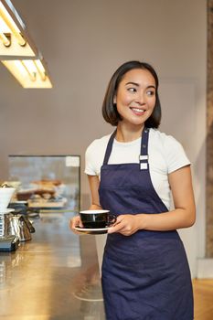 Portrait of asian smiling barista, girl serving coffee, standing near cafe counter in apron, preparing cappuccino for client.