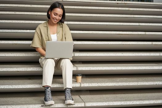 Portrait of young asian woman using her laptop, sitting outdoors on stairs. Happy smiling girl with personal computer.