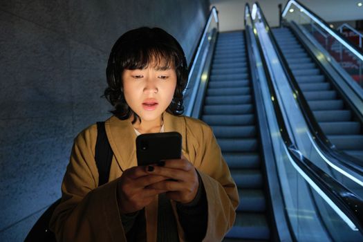 Portrait of asian girl looks shocked at mobile phone screen, standing on escalator, going down to underground or metro, commuting to work.