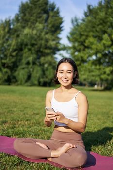 Portrait of korean girl meditating in park, follow yoga video lesson on smartphone app, sitting on fresh air and relaxing.