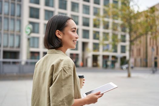 Asian girl walking on street, reading on tablet and drinking coffee outdoors.