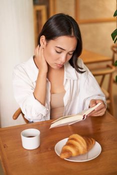 Asian girl reading book, looking sad, sitting in cafe and drinking coffee, has thoughtful sulking face.