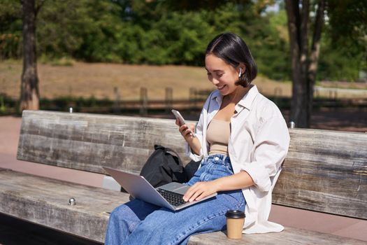 Young asian girl talking on mobile phone, using laptop, working online from park, sitting on bench on sunny day with computer.