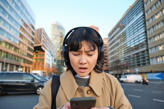 Portrait of young asian girl, student walks in city, listens music in headphones and uses mobile phone on streets, looks concerned at screen, reading bad news.