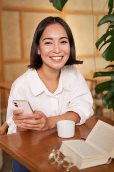 Portrait of korean girl in cafe, drinking coffee, using mobile phone and reading a book in cozy place.
