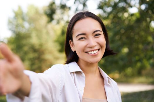 People and lifestyle. Happy asian woman takes selfie in park, photo on smartphone, smiling and looking joyful.