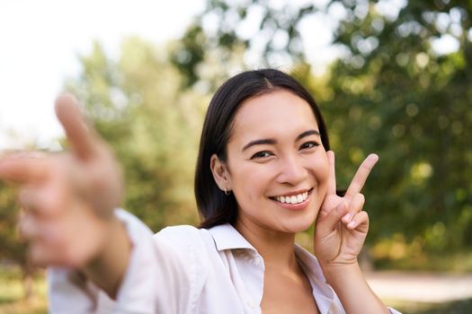 People and lifestyle. Happy asian woman takes selfie in park, photo on smartphone, smiling and looking joyful.