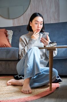 Vertical shot of young asian girl looks surprised at her mobile phone, reads amazing news, notification on smartphone app, sits at home on floor.