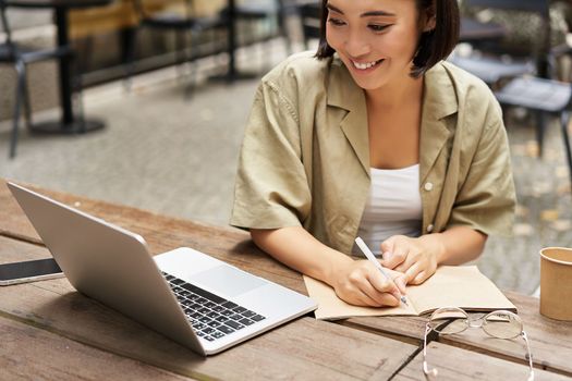 Portrait of young woman studying online, sitting with laptop, writing down, making notes and looking at computer screen, sitting in cafe outdoors.