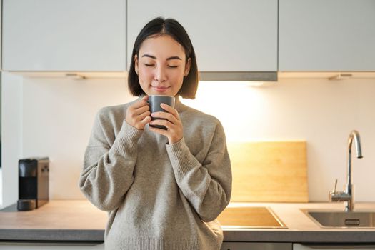 Portrait of smiling asian woman standing on her kitchen, drinking coffee and looking at camera, concept of cozy and comfortable home.