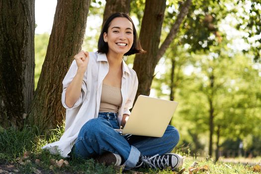 Enthusiastic young asian girl, sitting with laptop beside tree in green sunny park, celebrating, triumphing and smiling.