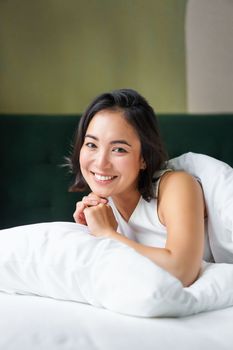 Vertical shot of smiling korean woman lying on pillow in her bed, spending morning in bedroom, waking up from sleep, looking happy.