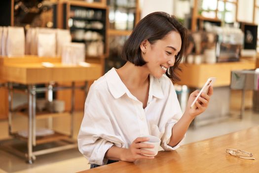 Beautiful asian girl, smiling and reading on smartphone, drinking coffee, sitting in cafe, looking at message on mobile phone.